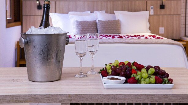 Un lit avec une literie blanche, une table de chevet avec une bouteille de champagne, de la glace, deux verres et une assiette de raisins et de fraises.