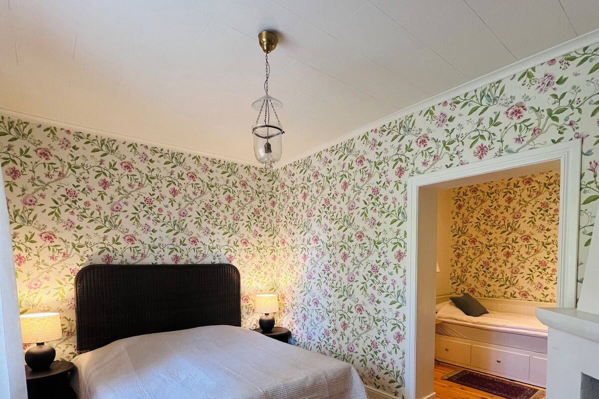 Ein Schlafzimmer mit einem Bett, einem Nachttisch, einer Lampe, Holzboden und floralem Wandpapier.