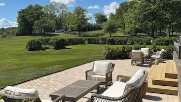 An outdoor patio with wicker furniture, a wooden table, and a view of a green lawn and trees.