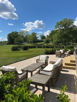 An outdoor patio with wicker furniture, a wooden table, and a view of a green lawn and trees.