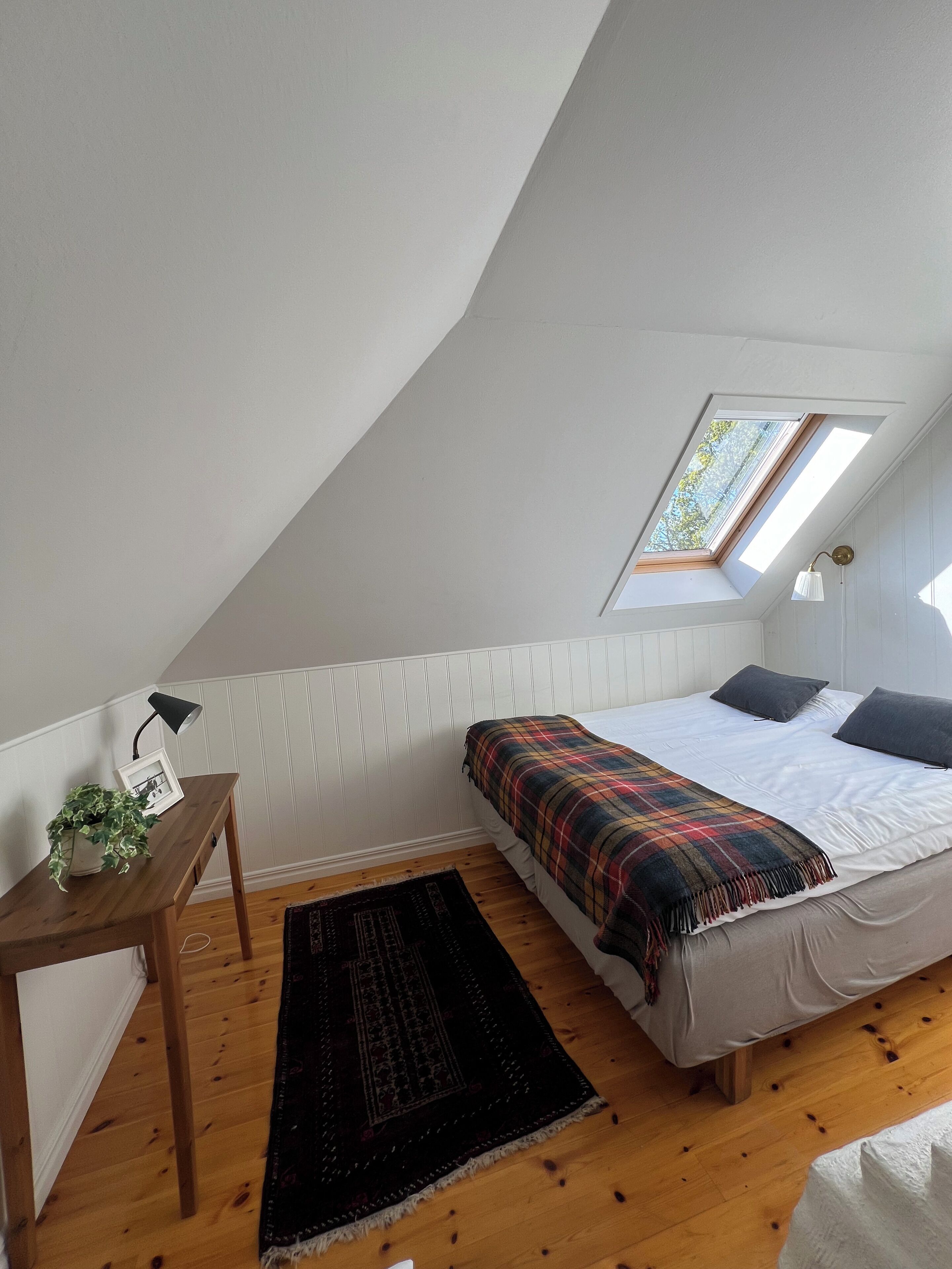 Ett sovrum med ett sluttande tak, ett takfönster, en säng med ett rutigt överkast, en sänglampa, ett litet bord med en växt och en mörk matta.