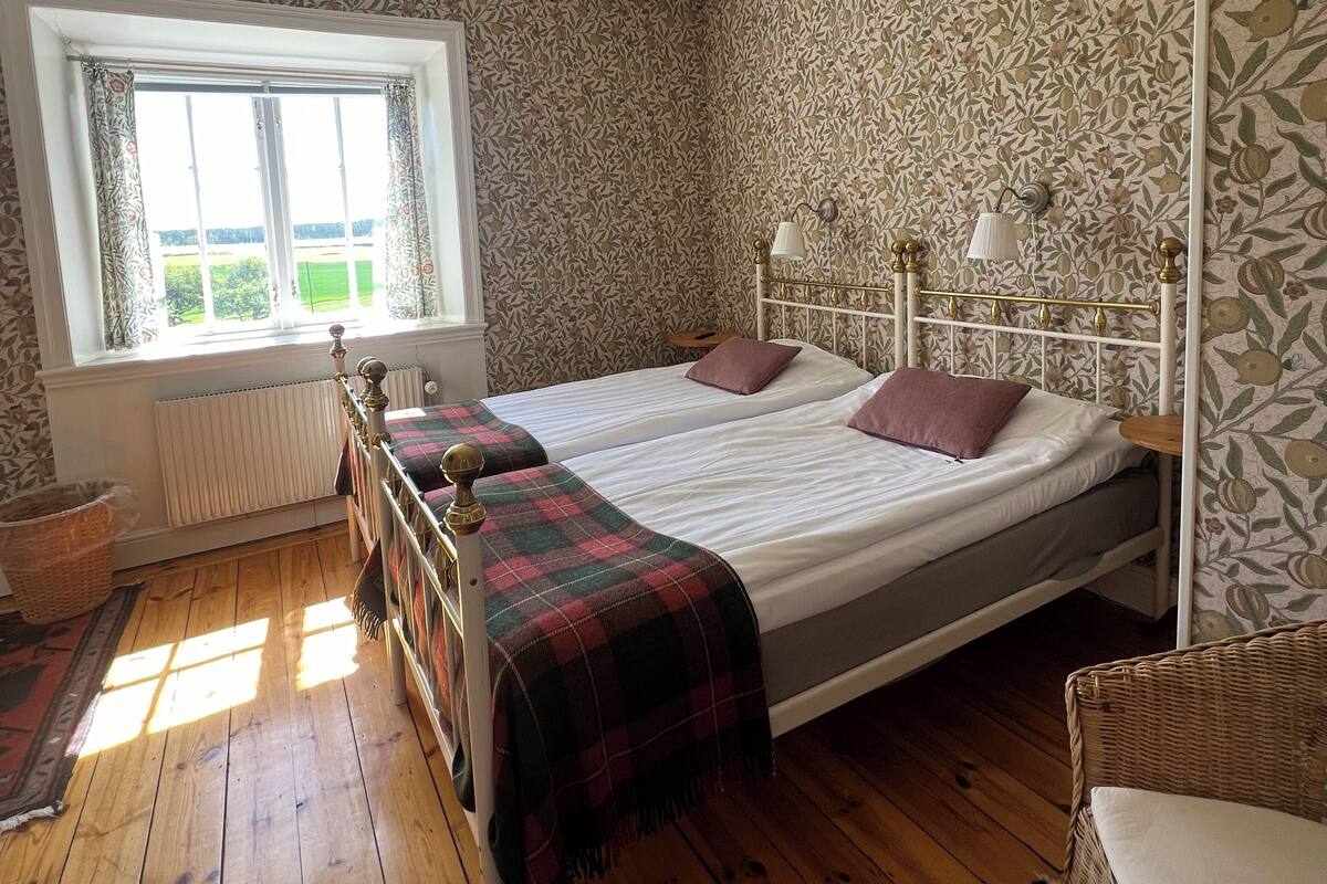 Ein Schlafzimmer mit zwei Betten, einem Fenster mit Vorhängen, einem Heizkörper und einem Wandpapier mit Muster.