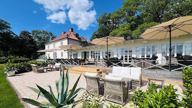 Een ruim terras buiten met rieten meubels en parasols, omgeven door weelderige begroeiing en een wit gebouw met een rood dak.