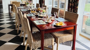 Een eetruimte met een geruit vloertje, rieten stoelen, een houten tafel gedekt voor een maaltijd en grote ramen met gordijnen.