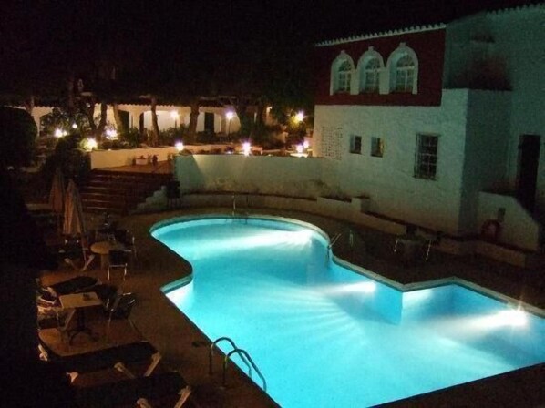A swimming pool with a curved edge, surrounded by lounge chairs and tables, and a building with arched windows in the background. - Hotel del Almirante (Es Castell)