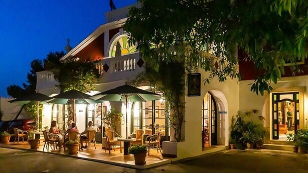 An outdoor dining area with tables and chairs, surrounded by potted plants and umbrellas, under a building with a balcony. - Hotel del Almirante (Es Castell)