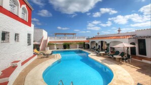 A swimming pool with lounge chairs and umbrellas, surrounded by a white building with red accents and a red-tiled roof. - Hotel del Almirante (Es Castell)