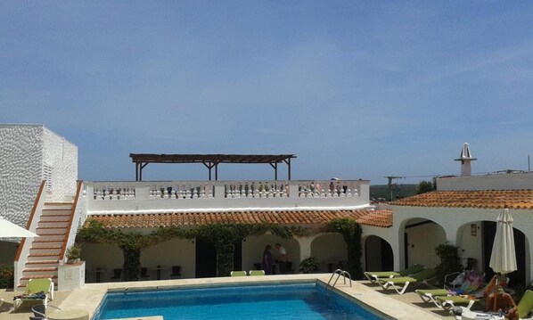 A rooftop pool area with a clear view of the sky, a covered terrace, and a building with a tiled roof.