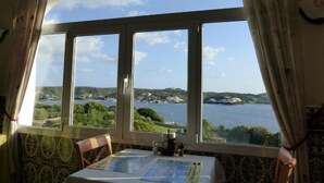 A dining area with a table and chairs, a view of a lake and hills, and a window with curtains. - Hotel del Almirante (Es Castell)