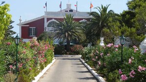 A building with a red facade and white trim, surrounded by lush greenery and pink flowers. - Hotel del Almirante (Es Castell)