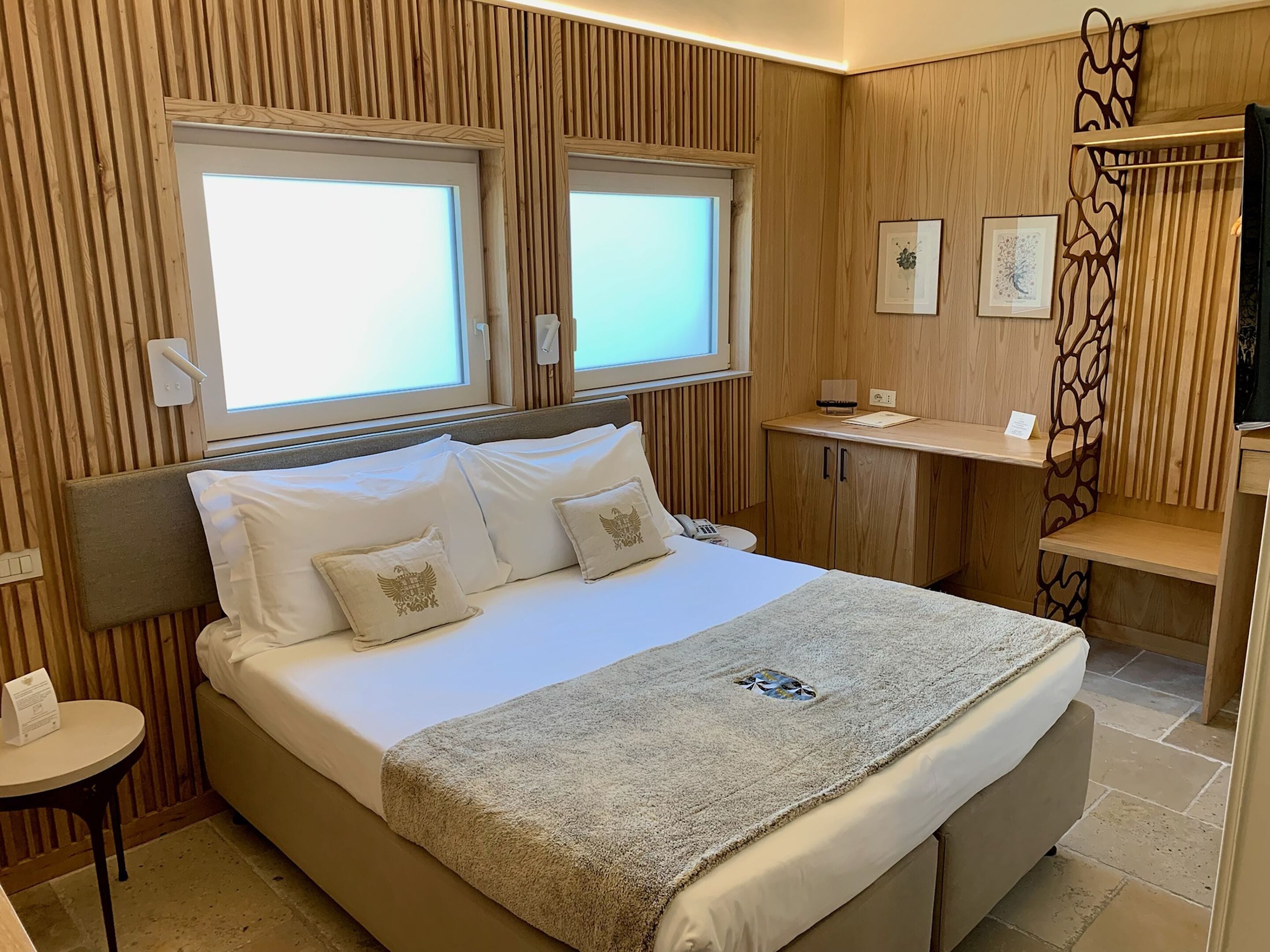 A modern hotel room with a large bed, two windows, a bedside table, a small desk, and a decorative wall panel.