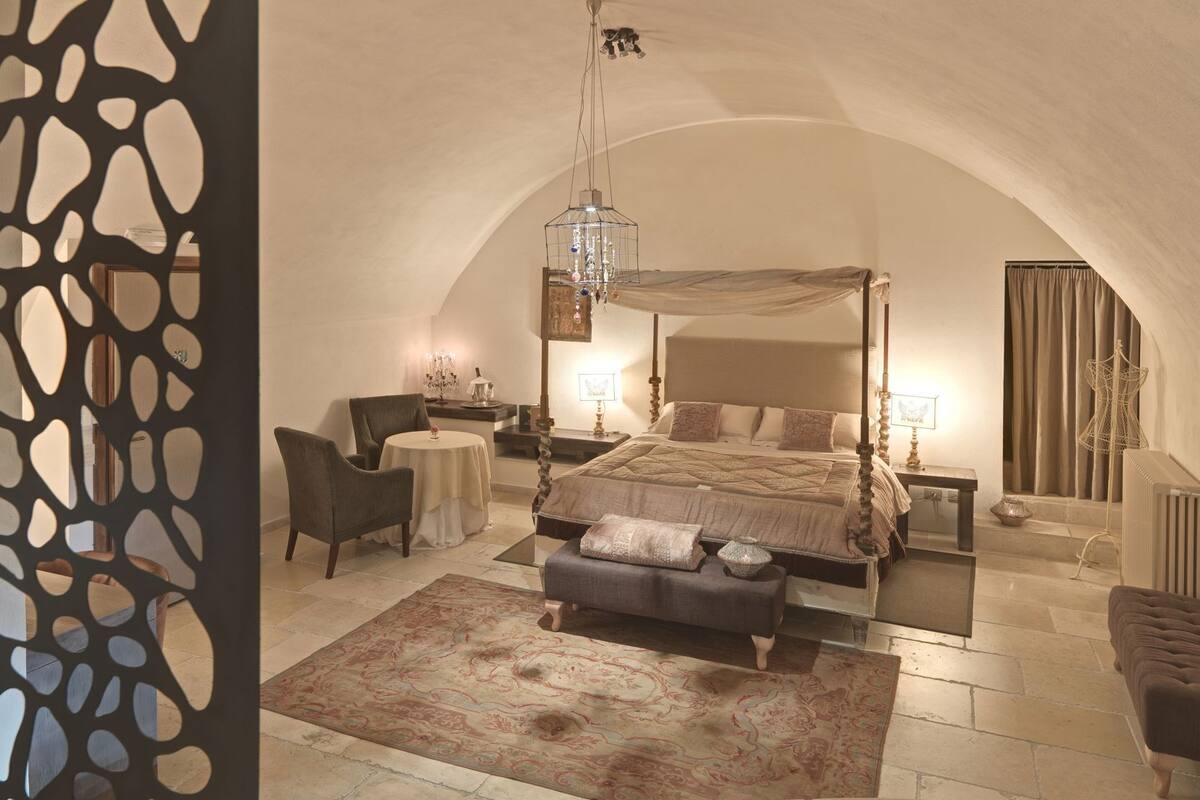 A bedroom with a four-poster bed, a chandelier, a round table, and a chair.