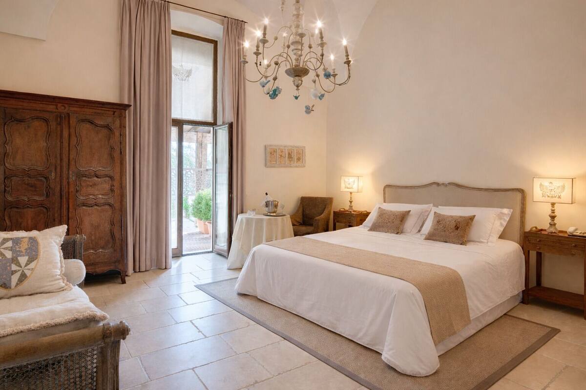 A spacious bedroom with a large bed, a chandelier, a wooden wardrobe, and a view through a glass door.