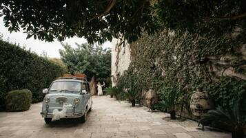 A vintage car parked on a paved driveway, surrounded by lush greenery and a stone wall.