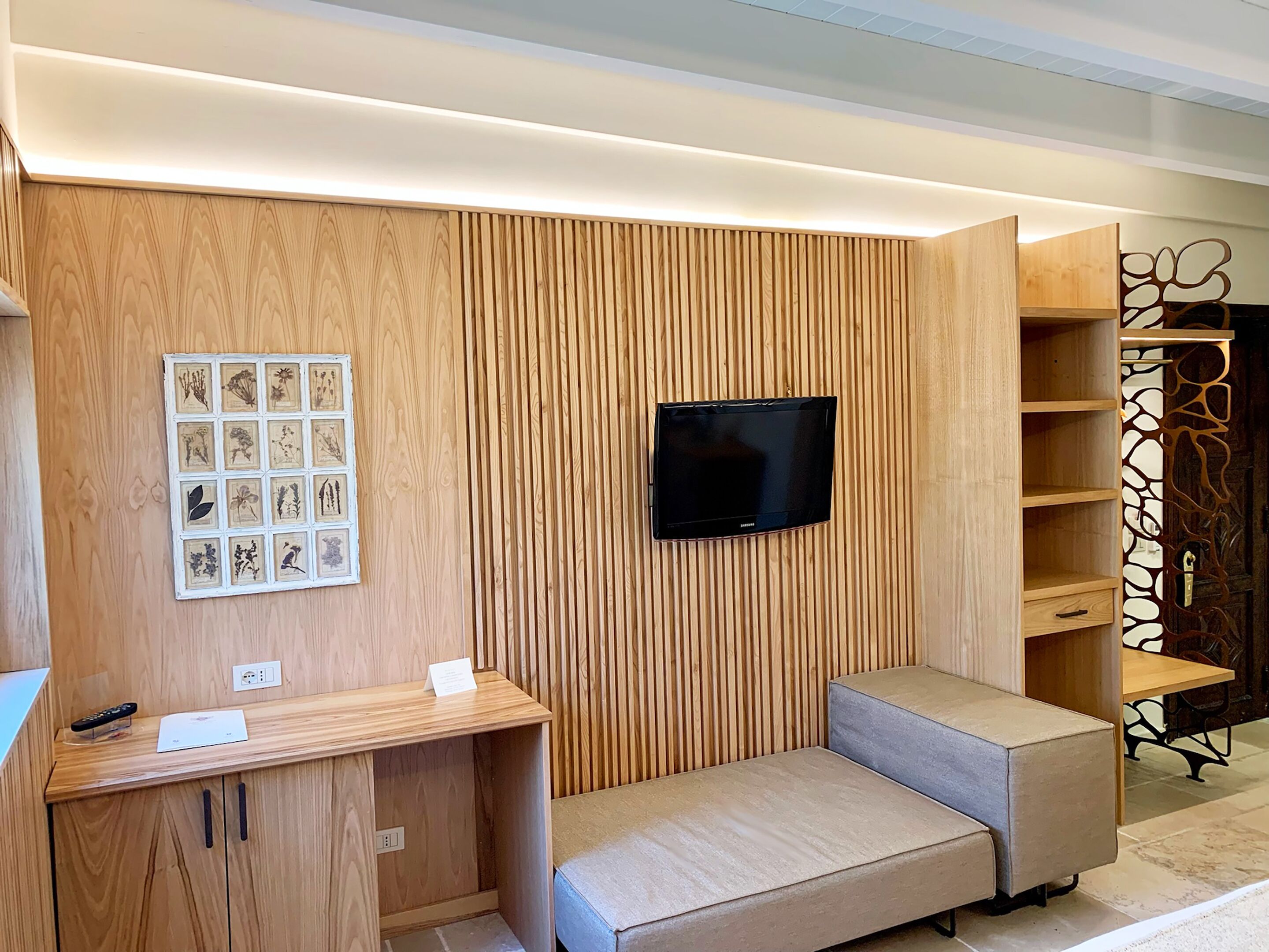 A modern living room with a wooden panel wall, a mounted TV, a small seating area, and a decorative shelf unit.