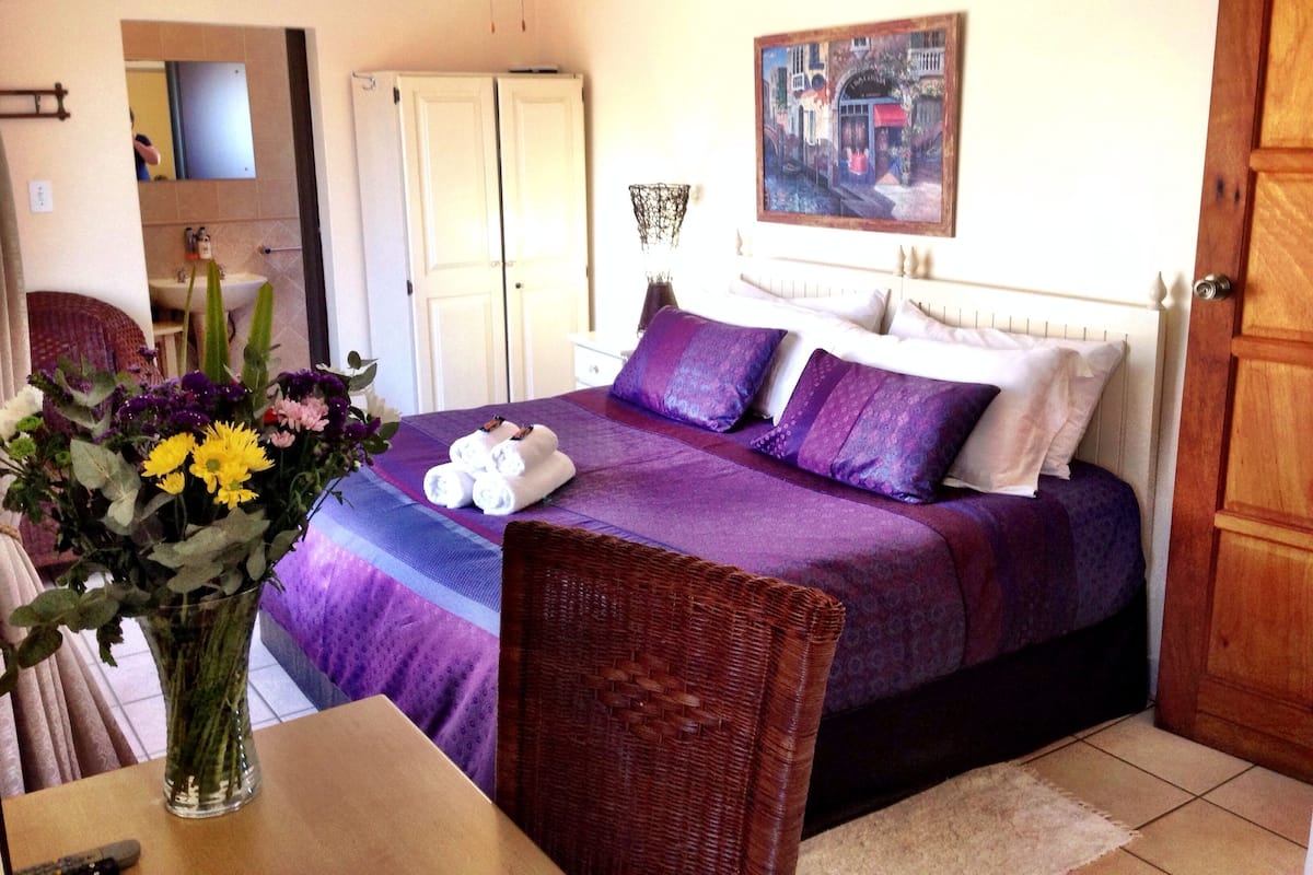 A bedroom with a bed, a wicker chair, a vase with flowers, and a painting on the wall.
