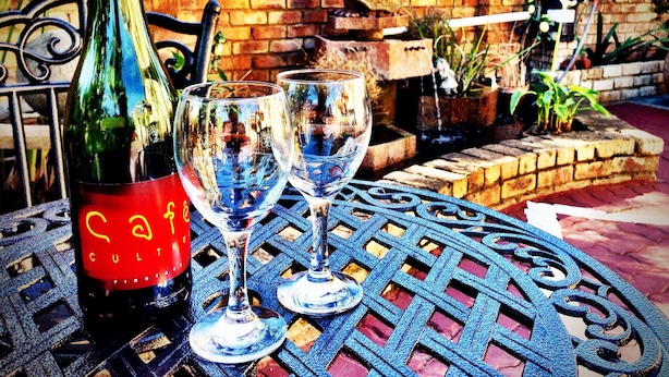 A bottle of wine and two glasses on a metal table with a brick wall in the background. - Pilgrims Delight (Centurion)