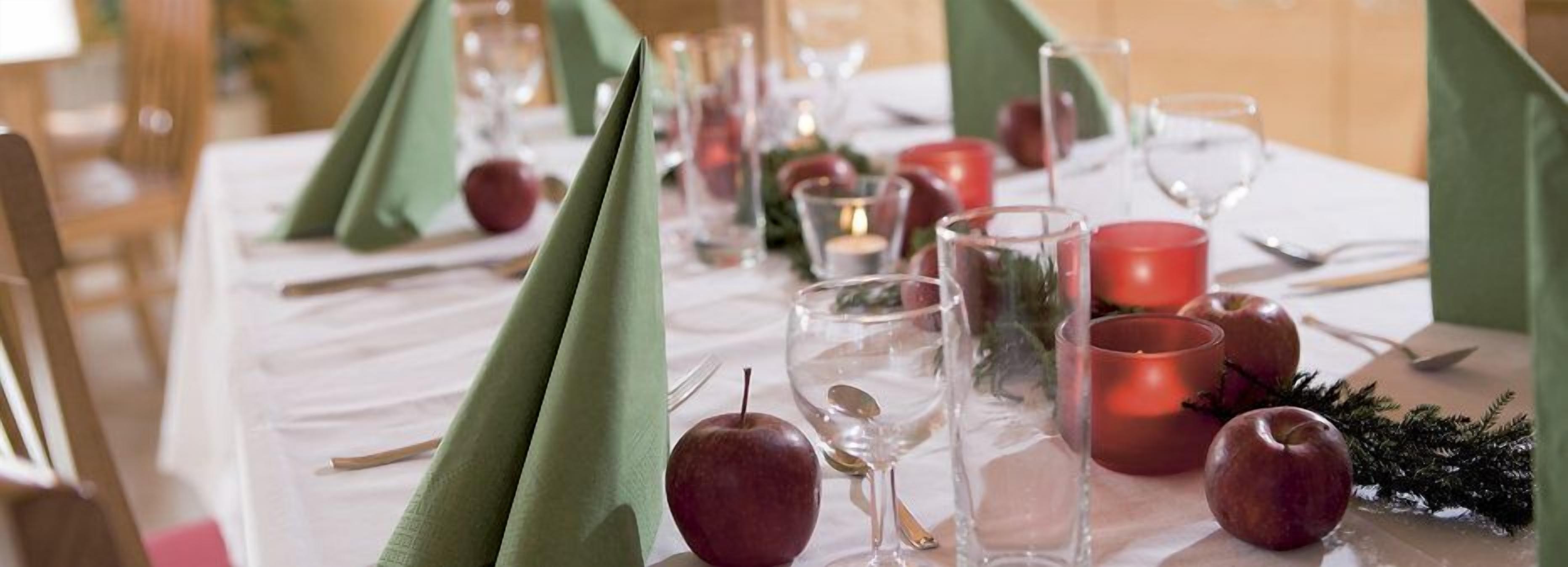Una mesa preparada para una comida, con servilletas verdes, manzanas rojas y vasos transparentes.