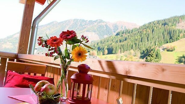 Una terraza de madera con una mesa y sillas, un florero con flores y un farol. Se observa un paisaje montañoso al fondo.