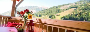 Una terraza de madera con una mesa y sillas, un florero con flores y un farol. Se observa un paisaje montañoso al fondo.