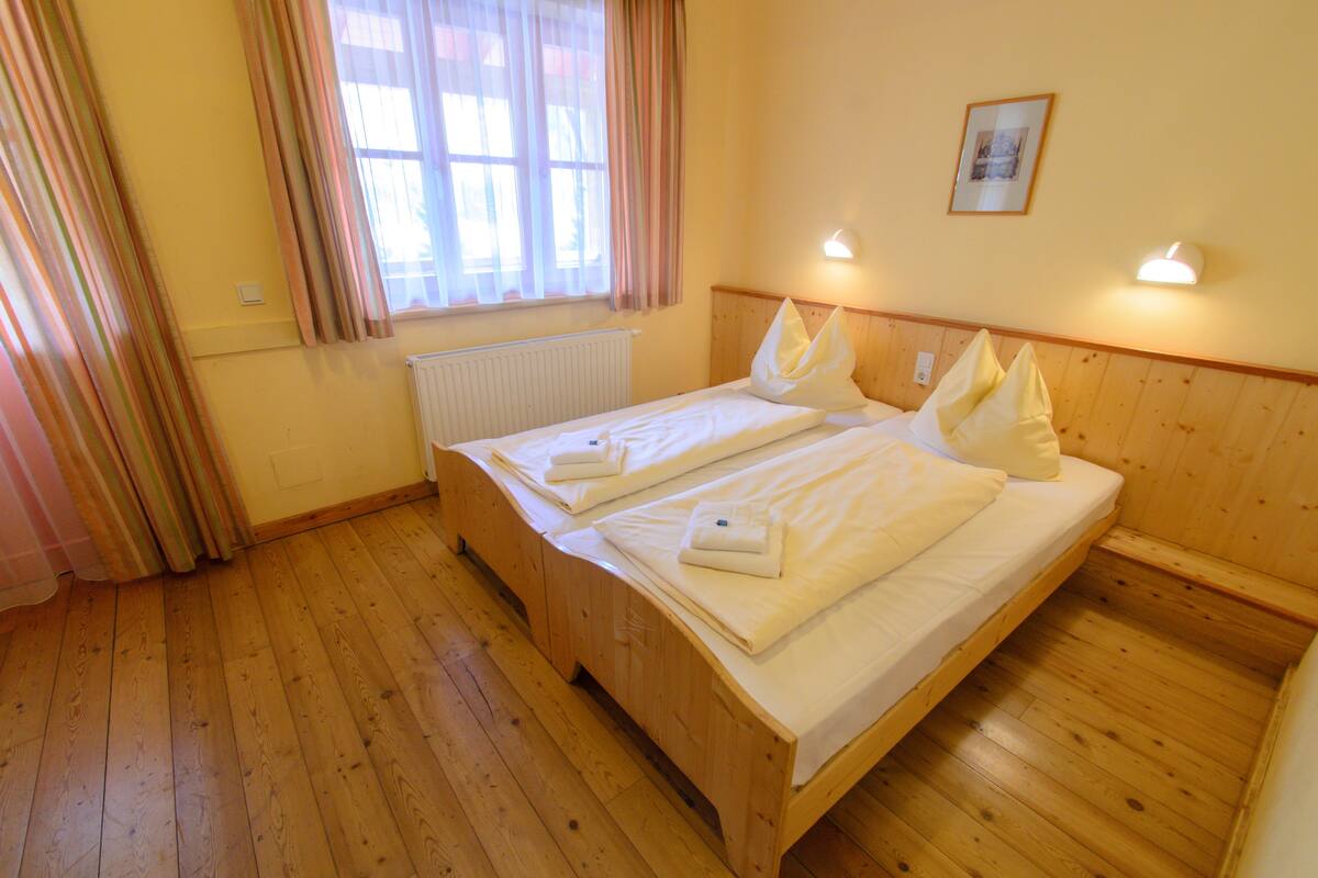 A double bed with white linens, a wooden headboard, a window with curtains, and a wall-mounted light fixture.