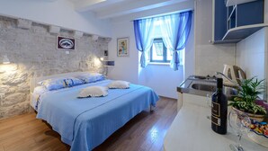 A bedroom with a bed, a window with blue curtains, a kitchen area with a sink, and a bottle of wine on the counter. - Studio Apartments Stradun (Dubrovnik)