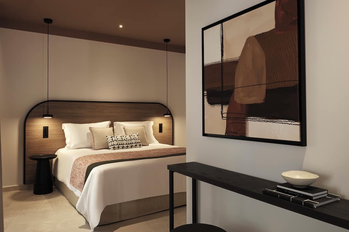 A modern bedroom with a wooden headboard, a bed with white bedding, a bedside table with a bowl, and a framed abstract painting on the wall.