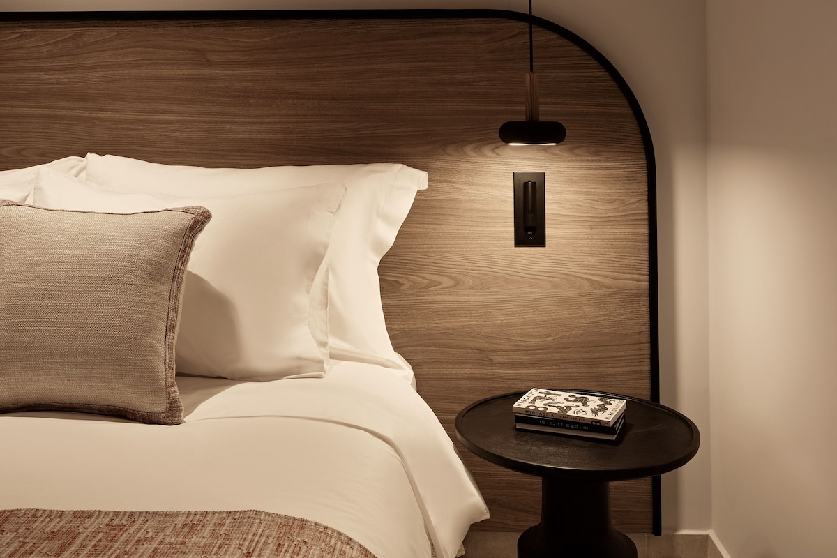 A neatly made bed with white linens and a brown throw pillow, a round side table with a book, and a pendant light fixture above the bed.