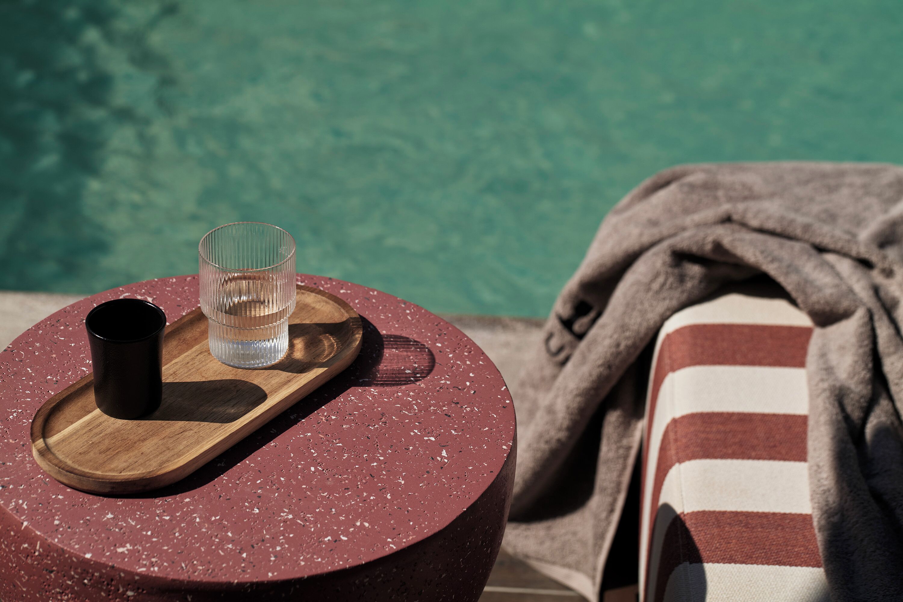 Una mesa redonda con una bandeja de madera que sostiene un vaso de agua y una taza negra, una toalla a rayas y vistas a la piscina.