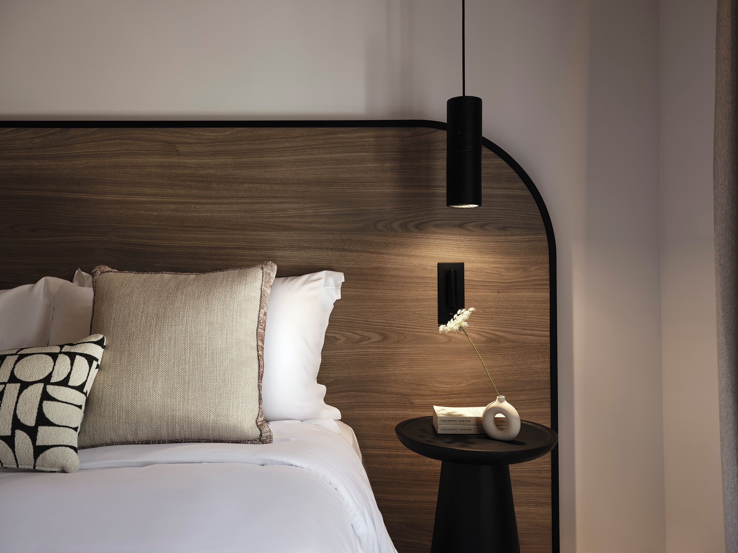 A modern bedroom with a wooden headboard, white bedding, and a small side table with a vase and a book.