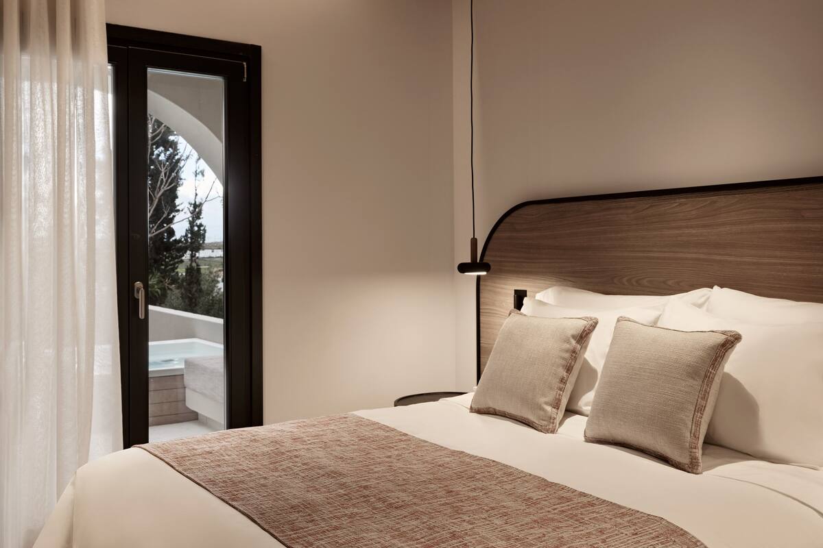 A modern hotel room with a large bed, a wooden headboard, and a view of the outdoors through a glass door.
