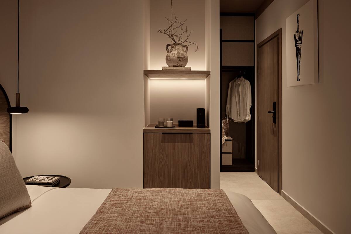 A modern bedroom with a wooden bedside table, a bedside lamp, a small shelf with decorative items, and a hanging robe.