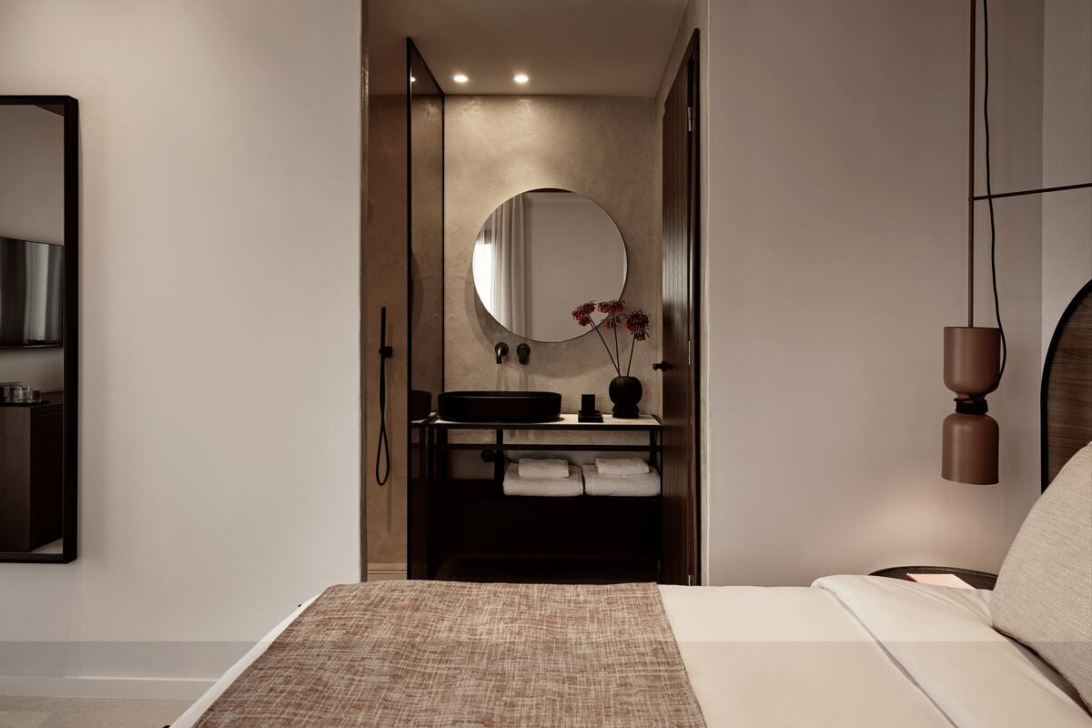A modern bedroom with a large bed, a bedside table, a mirror, and a bathroom with a sink and towel rack.