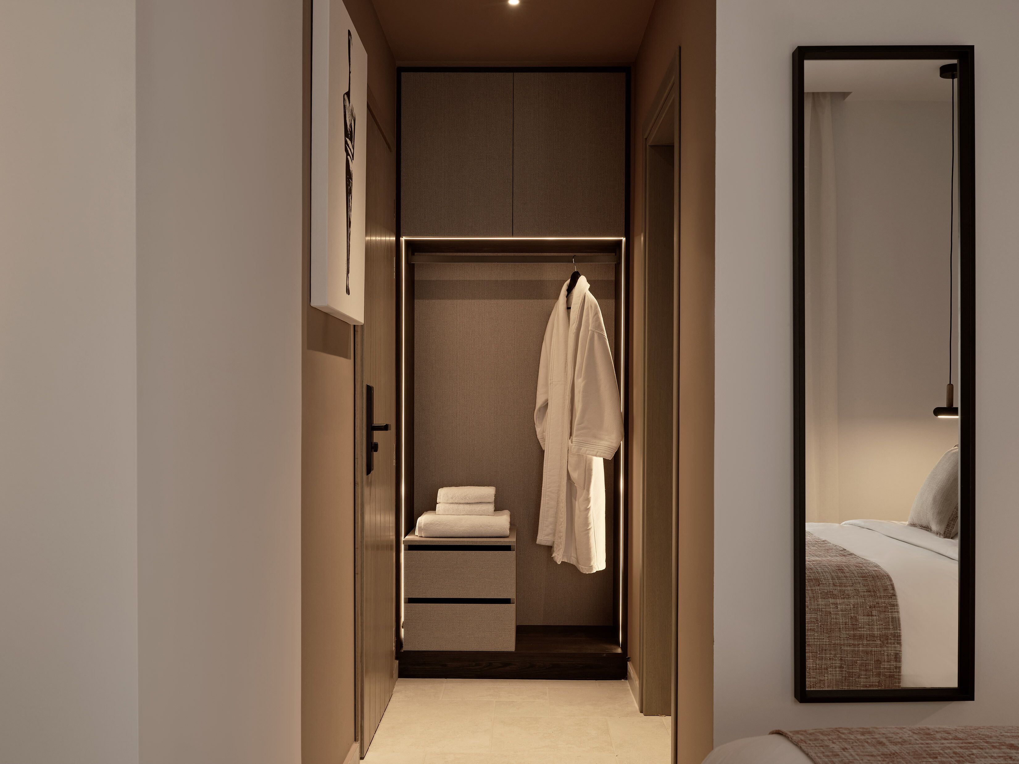 A modern hotel room with a walk-in closet, a bed with a textured blanket, and a mirror reflecting the room.