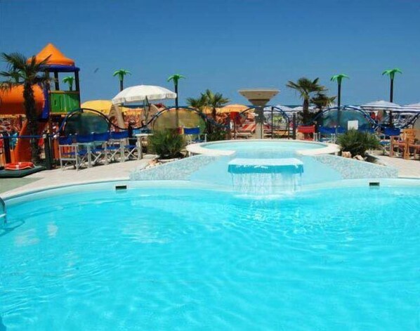 Una piscina con una fuente de agua, rodeada de palmeras y sombrillas de playa.