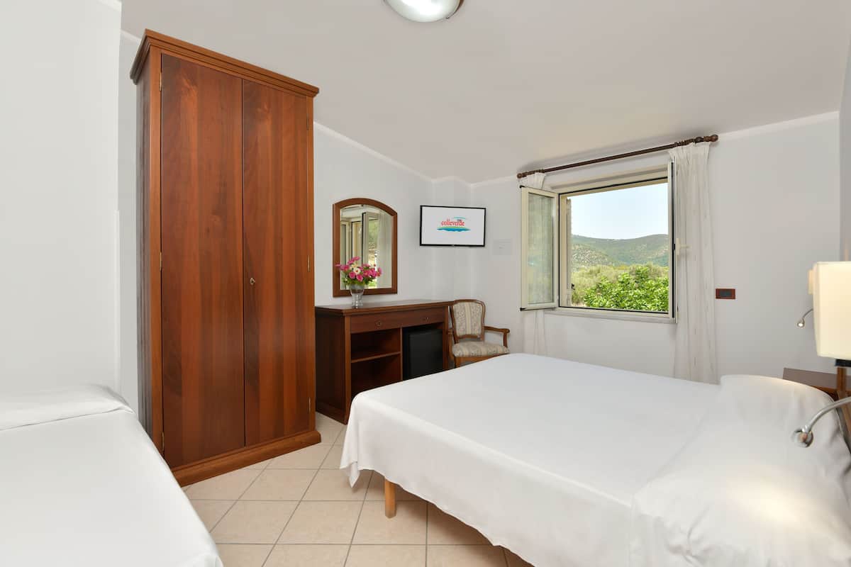 Ein Hotelzimmer mit zwei Betten, einem Holzschrank, einem Schreibtisch mit Stuhl, einem Fenster mit Blick auf Grünflächen und einem gerahmten Bild an der Wand.