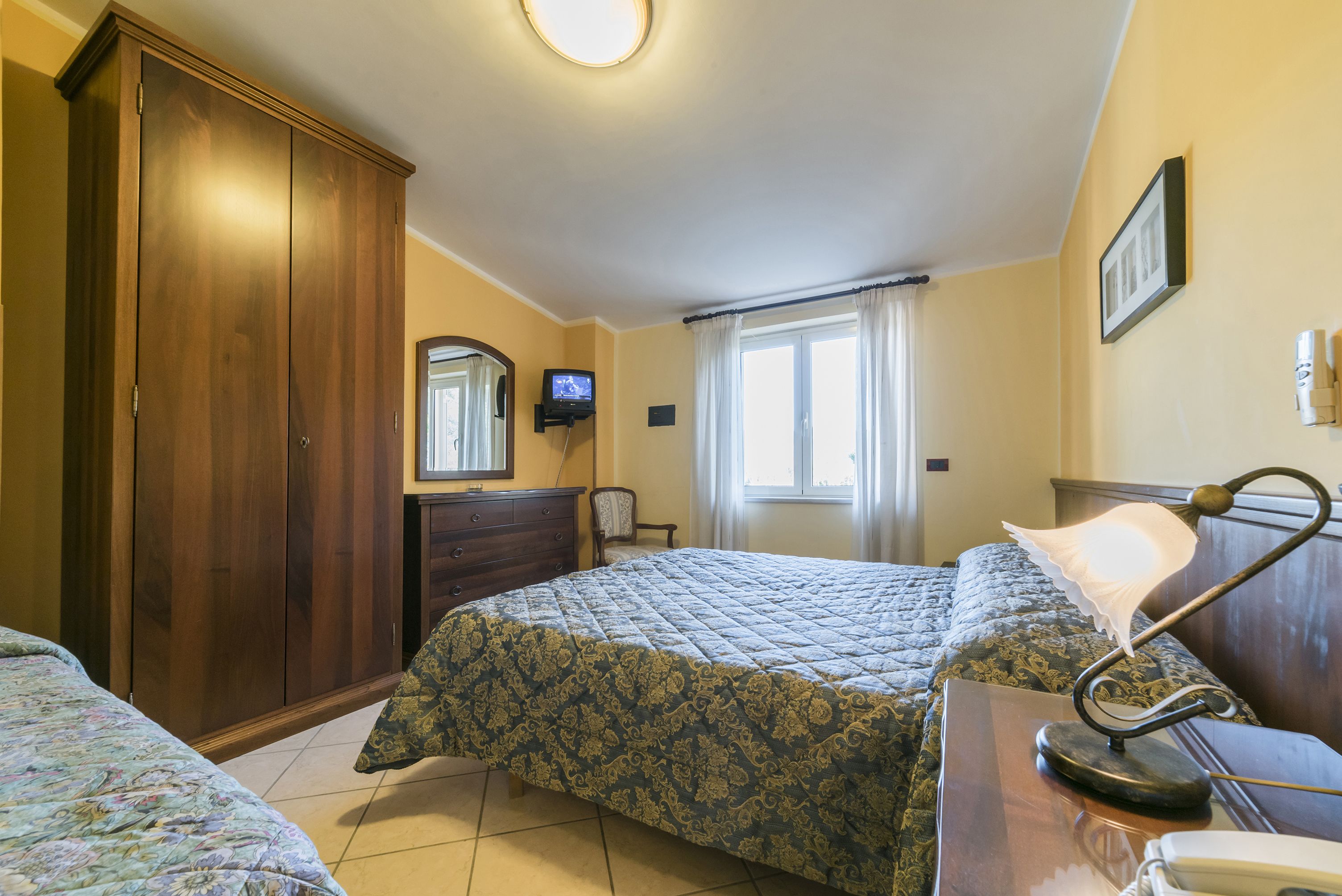 Ein Schlafzimmer mit zwei Betten, einem Kleiderschrank, einem Schreibtisch mit Lampe, einem Fernseher und einem Fenster mit Vorhängen.