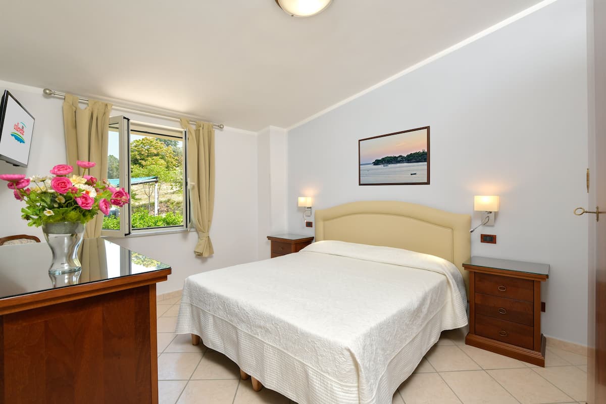 Ein Schlafzimmer mit Bett, Nachttischen, Schreibtisch, Stuhl, Fenster mit Vorhängen, einer Vase mit Blumen und einem Bild an der Wand.