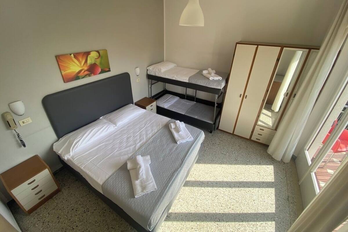 Ein Hotelzimmer mit einem Einzelbett, einem Etagenbett, einem Kleiderschrank, einem Schreibtisch mit Stuhl, einem Nachttisch, einer Lampe und einem Bild an der Wand.