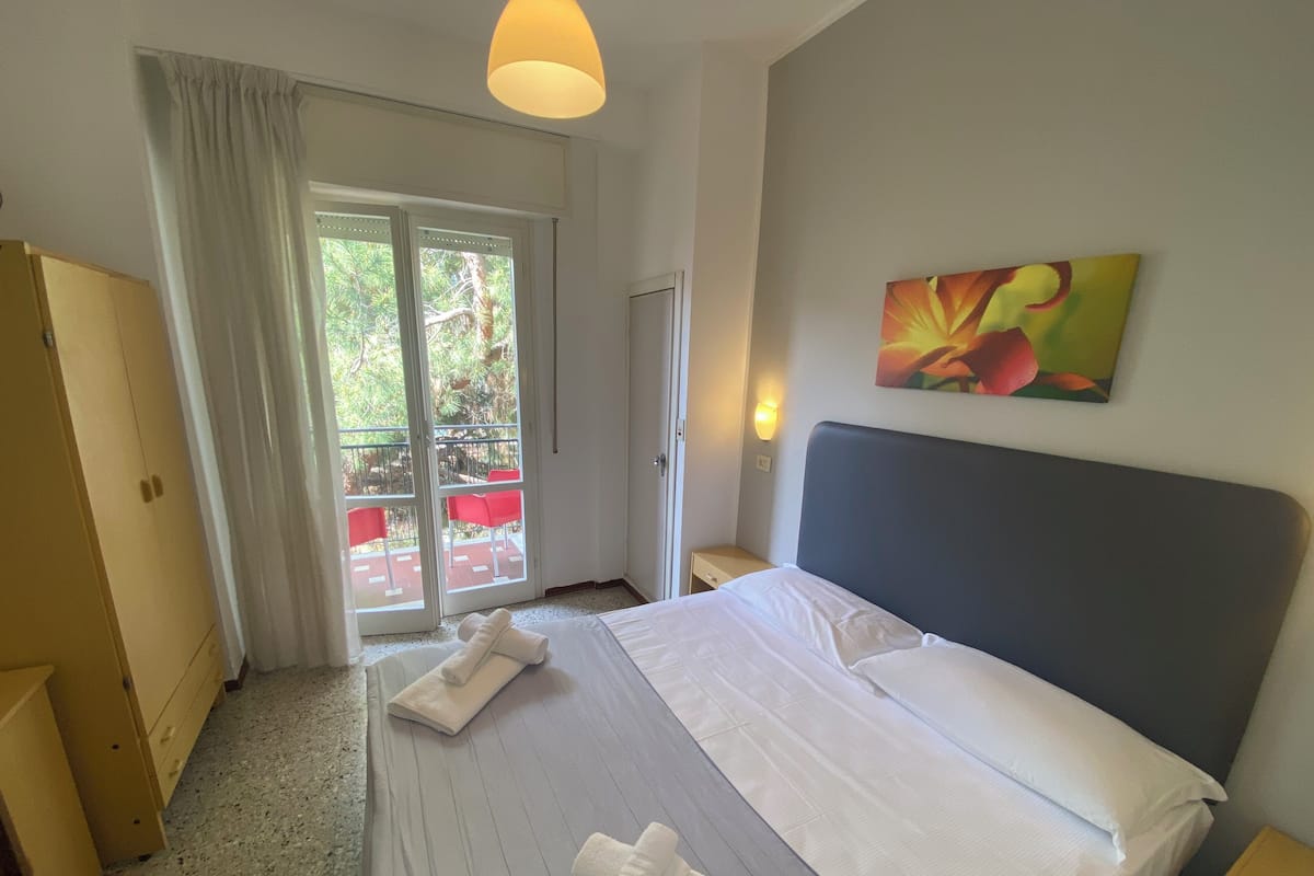 Ein Hotelzimmer mit einem Bett, einem Nachttisch, einem Gemälde und einer Glasschiebetür zu einem Außenbereich mit roten Stühlen.