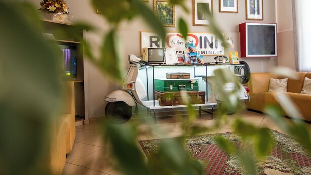 Ein Wohnzimmer mit einem Oldtimer-Roller, einem grünen Koffer und einer roten Kiste auf einem Tisch. An der Wand hängen gerahmte Bilder, und im Hintergrund steht ein Sofa.