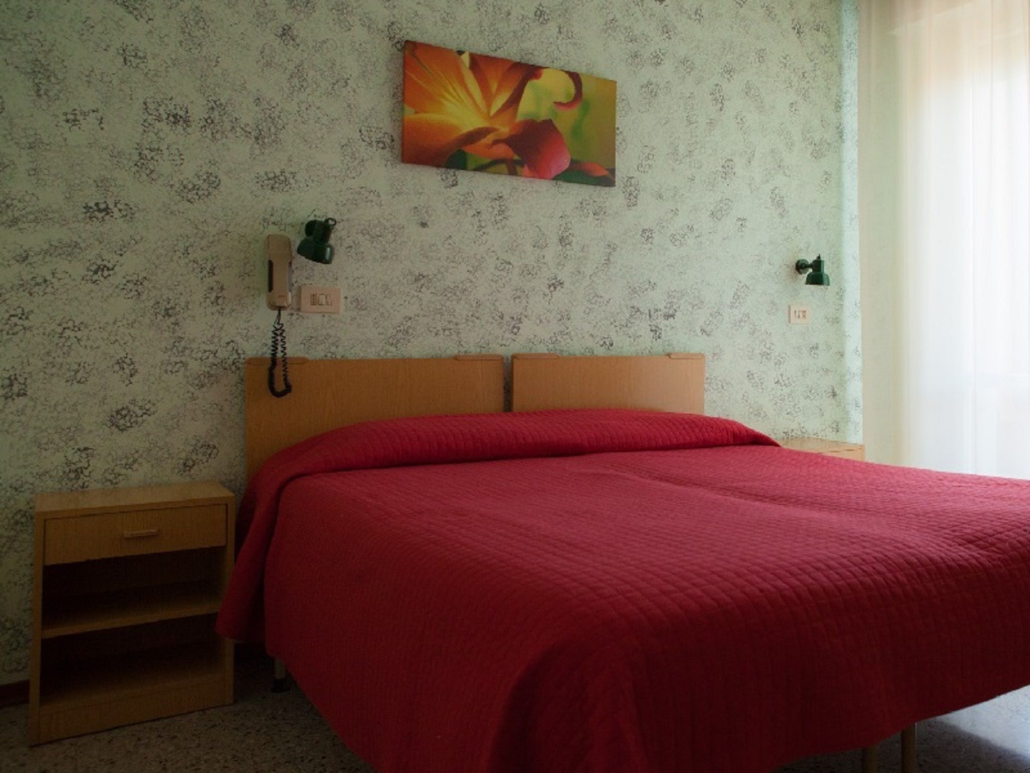 Un dormitorio con cabecera de madera, colcha roja, mesita de noche, teléfono y lámpara de pared.