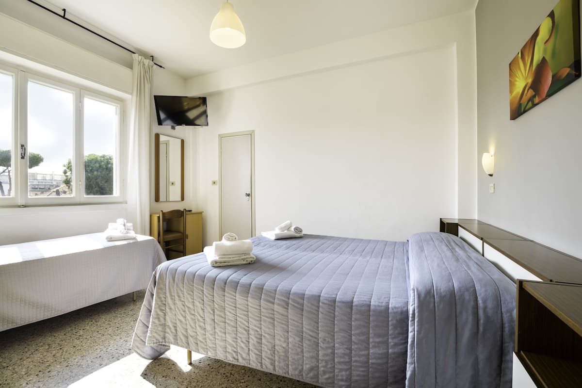 Ein Hotelzimmer mit zwei Betten, einem großen Fenster mit Vorhängen, einem Flachbildfernseher, einem Bild an der Wand und einem Nachttisch.