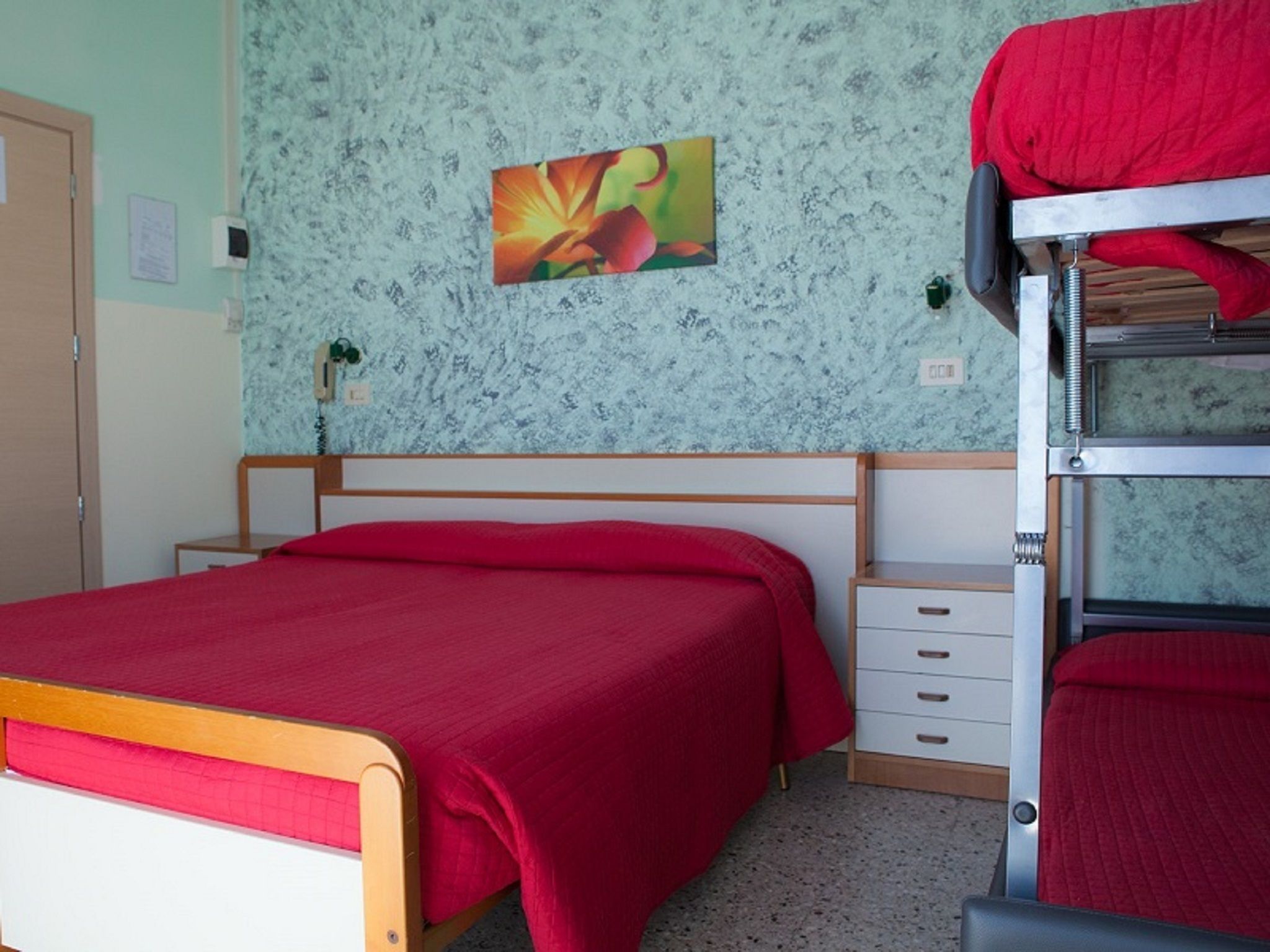 A bunk bed room with a red bedspread, a small bedside table with drawers, and a floral-patterned wall.