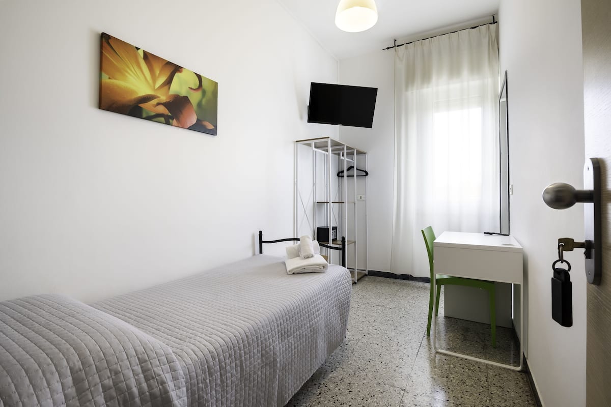 Ein Schlafzimmer mit Bett, Schreibtisch, Stuhl, Fernseher, einem Bild und einem Fenster mit Vorhängen.