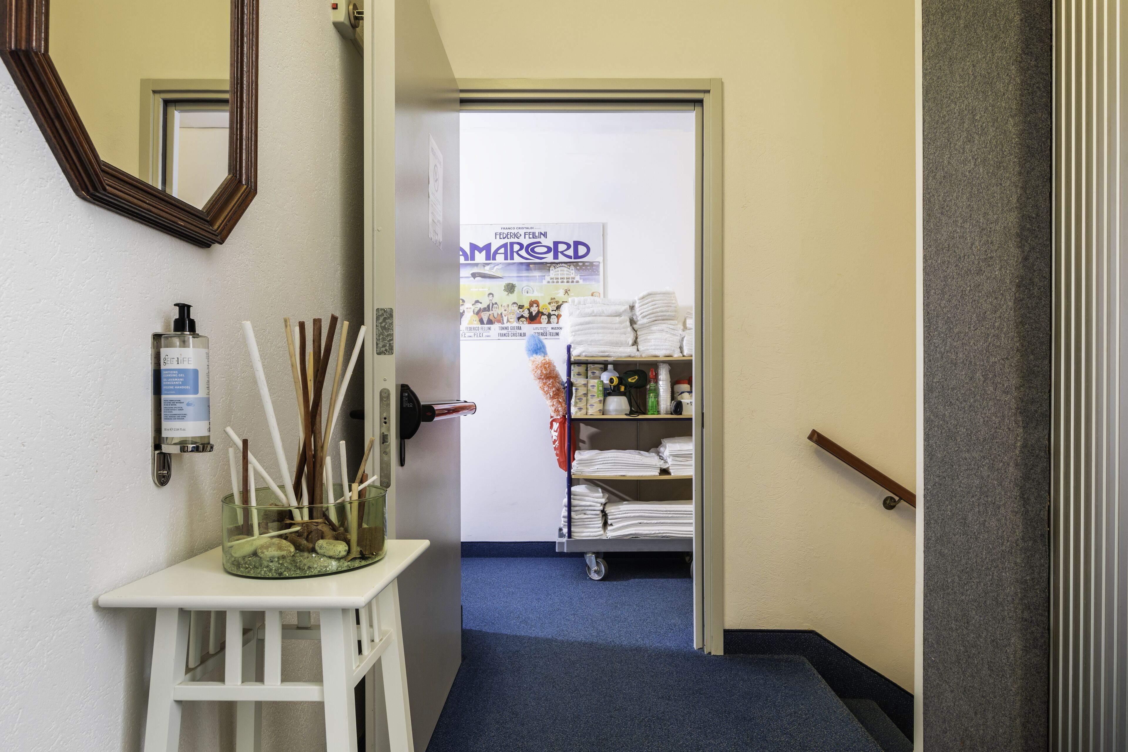 A hallway with a mirror, a small table with a bottle and straws, a poster, and a cart with towels.