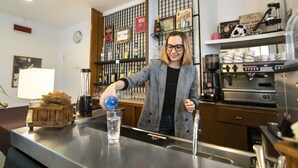 Ein Barista bereitet ein Getränk an der Theke zu, im Hintergrund sind Kaffeemaschinen und verschiedene Kaffeetassen und -becher auf Regalen zu sehen.