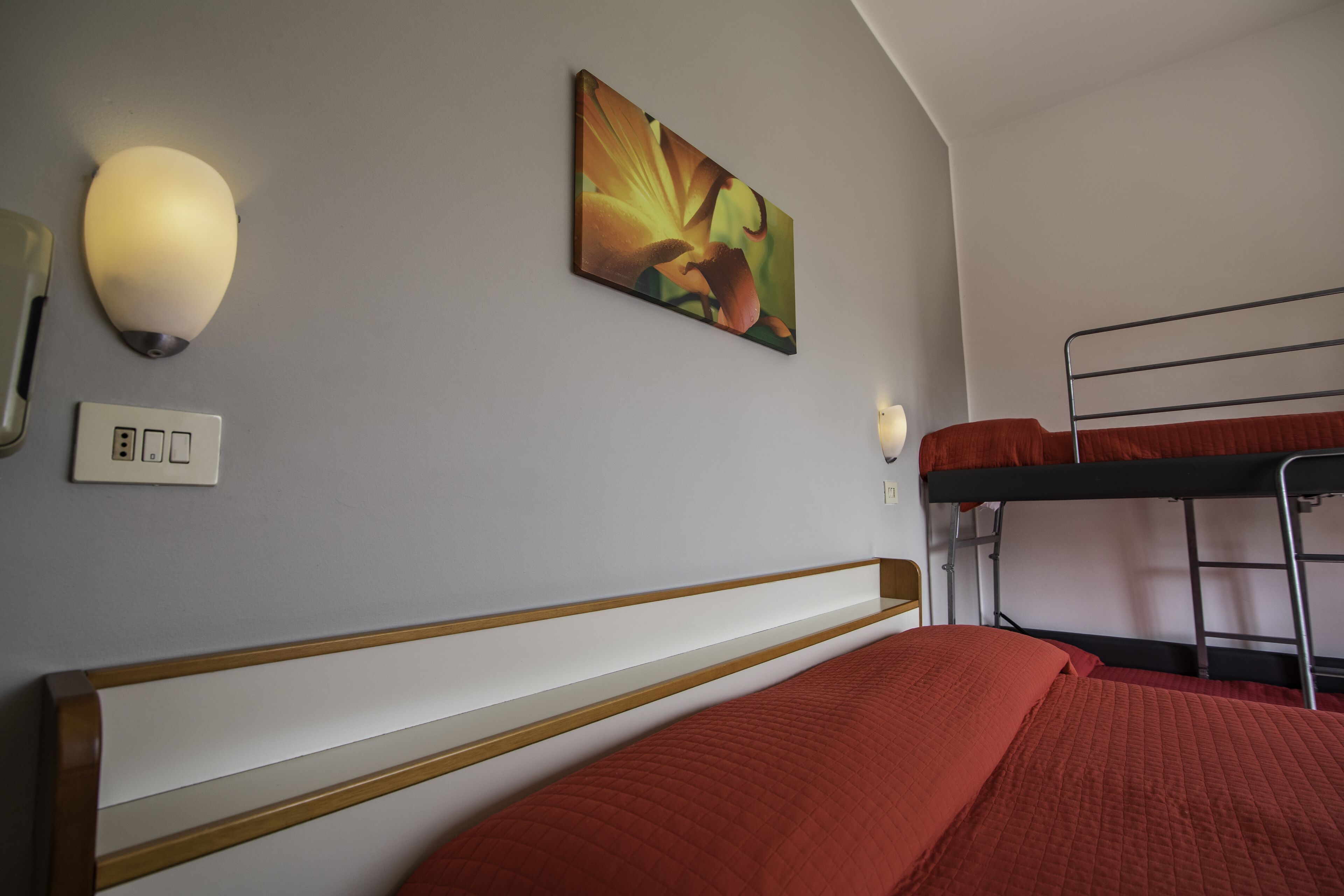 A bunk bed with red bedding, a wall-mounted light, and a painting.