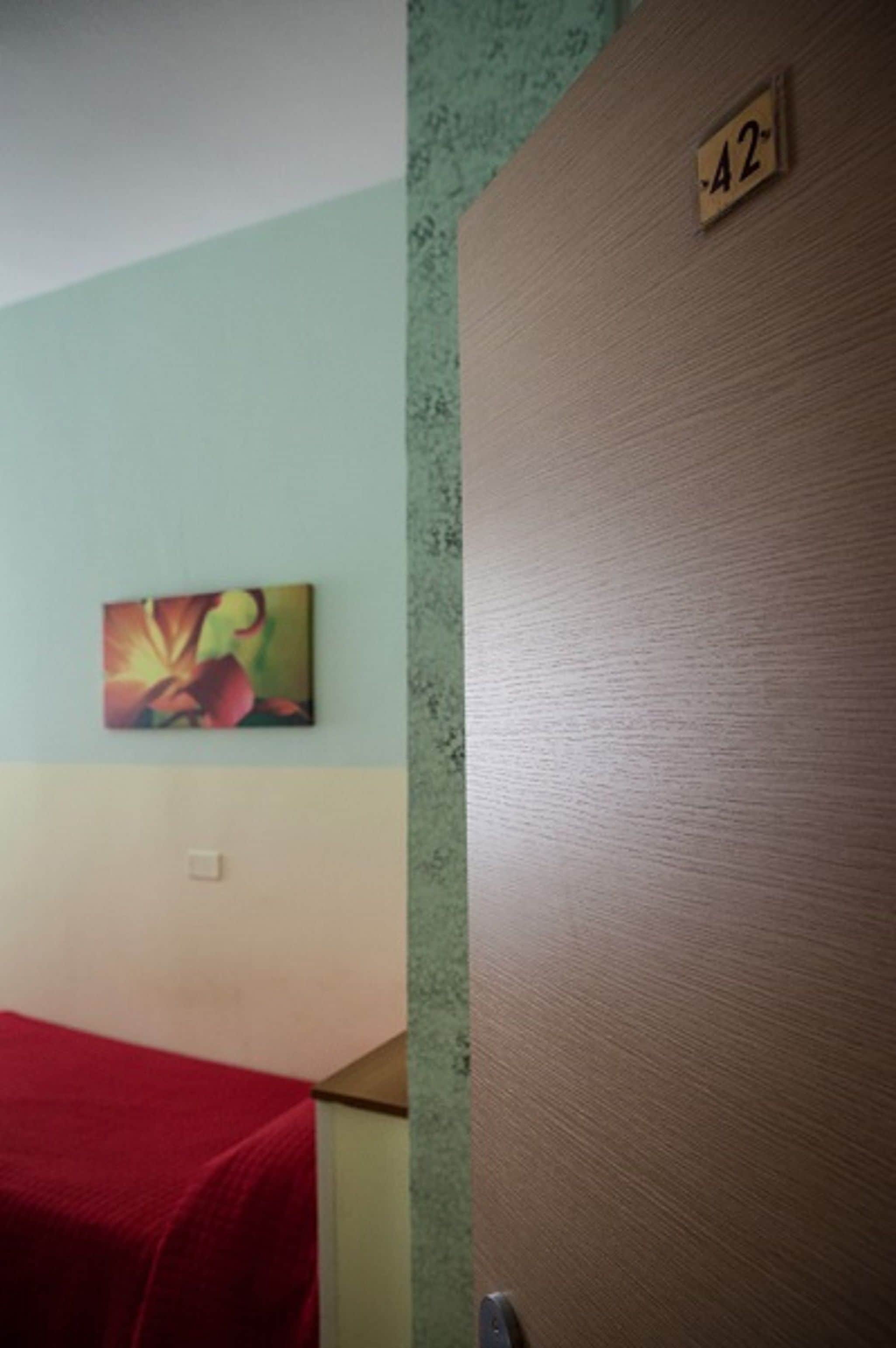 A room with a bed covered in a red blanket, a wall-mounted painting, and a door with a number plate.