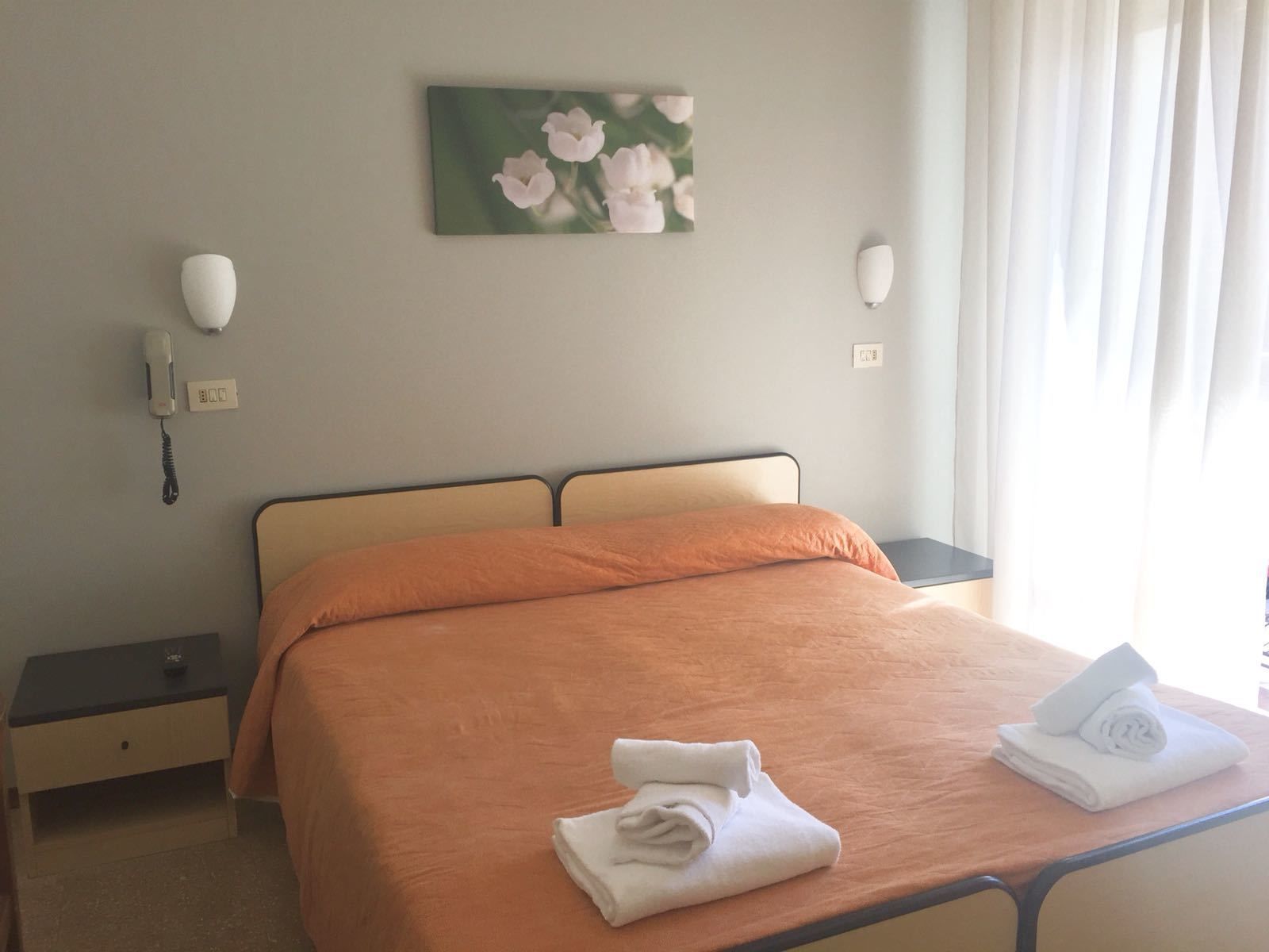 A hotel room with a bed, a nightstand, a telephone, a painting, and a window with curtains.